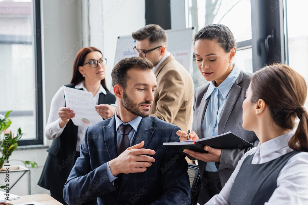 African american businesswoman holding notebook near colleagues in office African american businesswoman holding notebook near colleagues in office