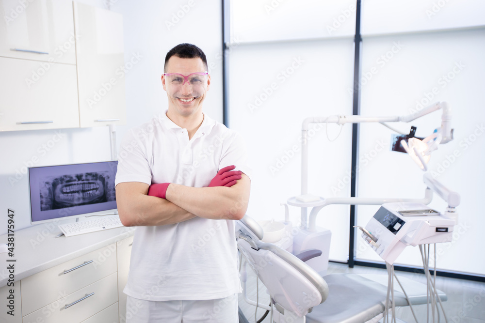 Portrait of a young smiling dentist in work glasses and in a white uniform. The man folded his arms and stands at the dental chair in the office