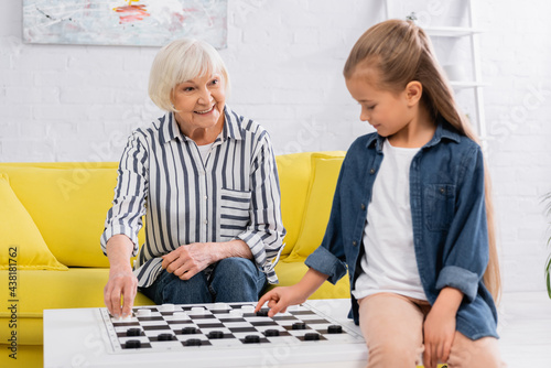Smiling elderly woman playing checkers game with blurred granddaughter