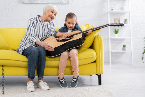 Smiling kid playing acoustic guitar near grandparent on sofa