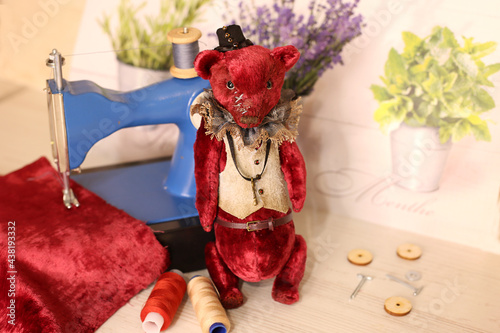 Red teddy bear stand next to a sewing machine, fabric and thread, selective focus