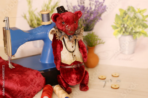 Red teddy bear stand next to a sewing machine, fabric and thread, selective focus