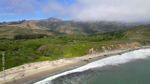 Aerial drone flying alongside ocean cliffs with greenery and mountains in the background.