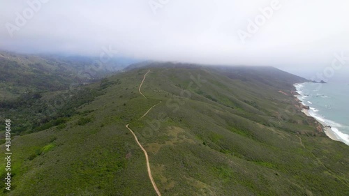 Aerial drone flying alongside ocean cliffs with greenery and mountains in the background.