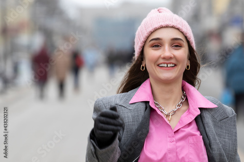 A positive young girl cheerfully walks along the city street waving her arms. A teenage girl with a gap between her white teeth walks around the city wearing a hat with gloves and an unbuttoned coat.