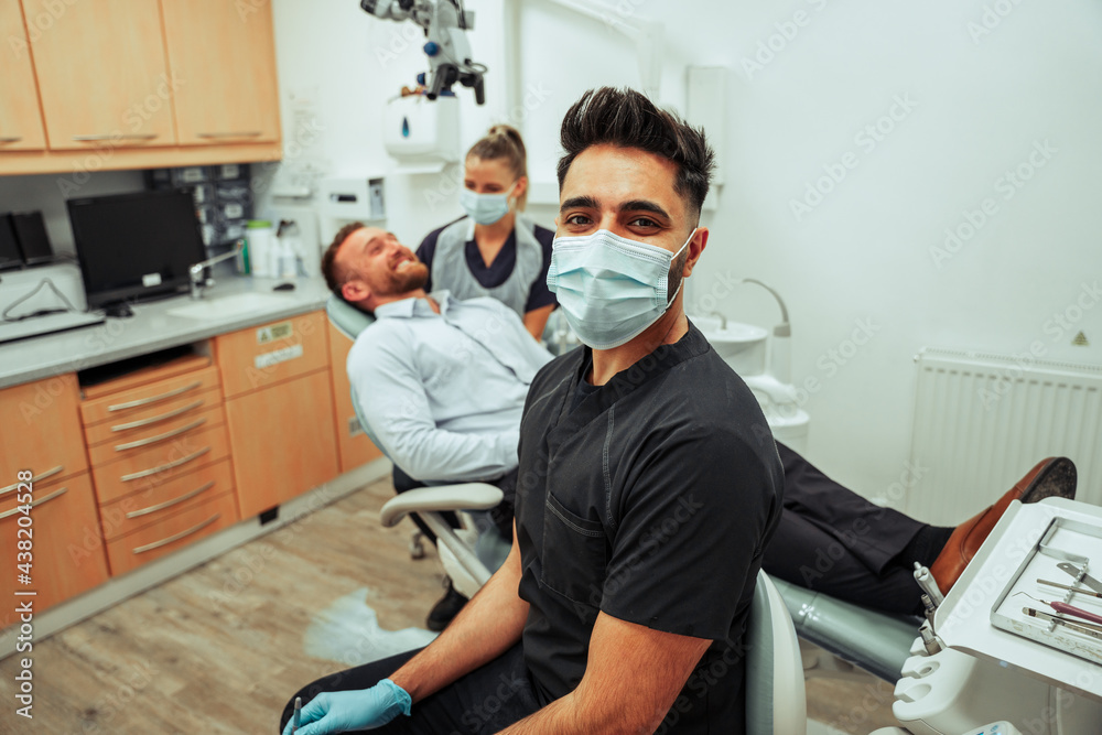 Caucasian male nurse sitting with surgical mask over seeing female dentist clean male client teeth
