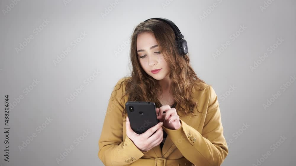 Attractive woman in earphones choose music in phone and dances standing on a white background. A young caucasian lady in yellow costume with smartphone in her hand is moving and enjoying the song