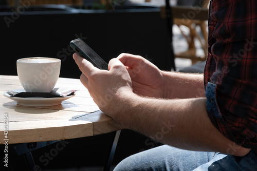 Close up of hands of a man writing something on his mobile phone. The hands rest on a wooden table on a terrace with a cup of coffee on it. Coffee break