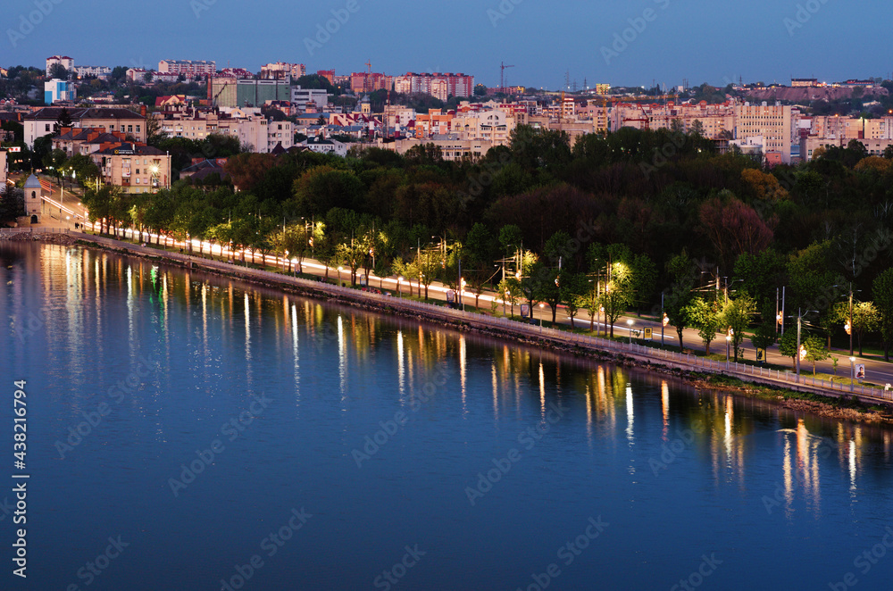 Fototapeta premium Ternopil, Ukraine-May, 11,2021:Night lights landscape of Ternopil. Motorway between city lake and park with green trees. Colorful light trails with motion blur effect. Romantic and peaceful scene