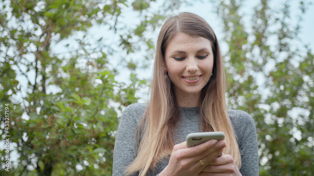 Happy woman typing by mobile phone outdoors. Cheerful girl with smartphone in park on a background. Smiling lady holding cellphone in hands outside. Sport Girl, internet technology, runner exercise Happy woman typing by mobile phone outdoors. Cheerful girl with smartphone in park on a background. Smiling lady holding cellphone in hands outside. Sport Girl, internet technology, runner exercise