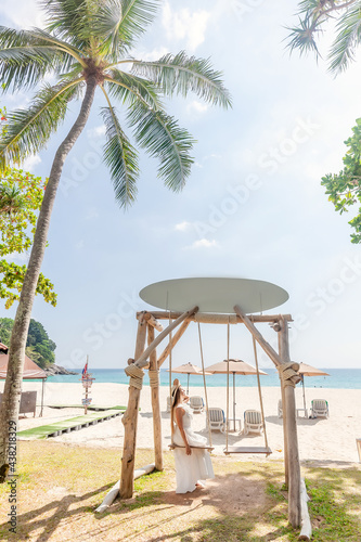 Travel Woman in White Dress and Straw Hat Sitting on Wooden Swing, Tropical Palms and Sandy Beach with Blue Sea on Background. Female Tourist Leisure in Resort on Phuket Island, Thailand © TravelMedia