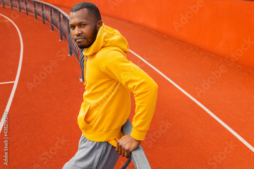 Portrait of a smiling afro american sportsman looking at camera, wearing bright yellow sportswear resting after hard work out and leaning on a wall outdoors at stadium © shunevich
