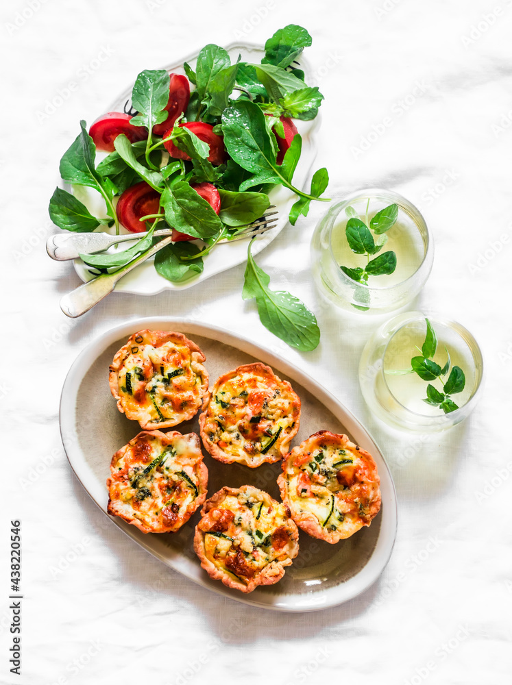 Aperitif table - mini quiche with salmon and zucchini, fresh tomato arugula salad and mint cocktail on a light background, top view