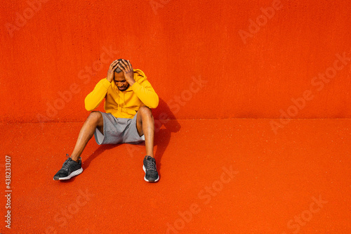 African America athlete sportsman with yellow sportswear sitting isolated on colorful red background in a stadium. Serious tired black male look down, holding head with both hands, losing game