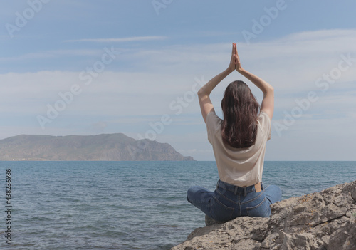 A girl sits by the sea and folds her arms namaste over her head. Meditating and relaxing