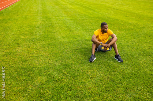 Young pensive Afro American sportive male sitting with smartphone on grass in the middle of stadium, relaxing after workout training, football soccer match. Copy space. Healthy sport lifestyle concept