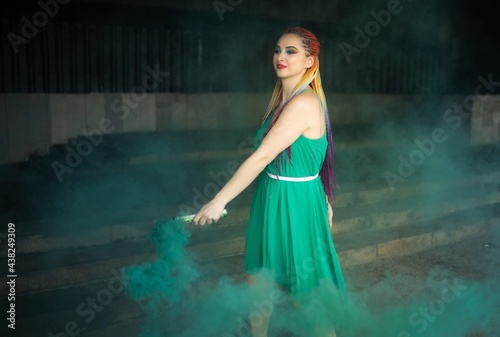 Cute girl in a green dress with African braids and colorful makeup posing with bright green smoke on the streets of a warm spring city