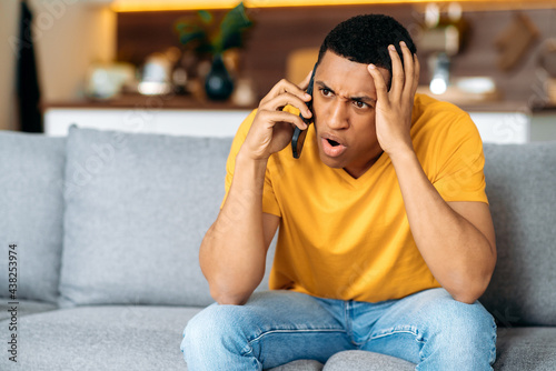 Confused hispanic guy sitting on the couch in the living room, dressed in casual shirt, bewildered, talking on the phone, angry, annoyed, have unpleasant telephone conversation
