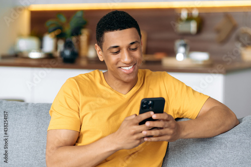 Joyful latino attractive guy using his smartphone while sitting on sofa in stylish clothes, browsing internet and social networks, texting with friends or family, found out good news, smiles happily