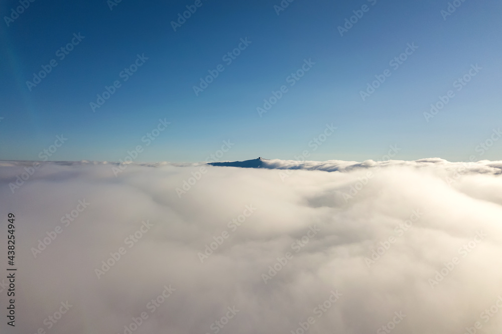 Aerial view from above of white puffy clouds and distant mountain top in bright sunny day.
