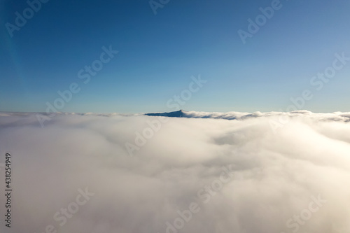 Aerial view from above of white puffy clouds and distant mountain top in bright sunny day. © bilanol