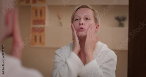 Blond hair woman looking in the mirror, applies cream on her face and neck