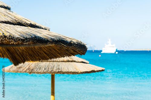  Selective focus  Stunning view of a thatch umbrella in the foreground and a luxury yacht sailing on a beautiful turquoise sea in the background. Sardinia  Italy.