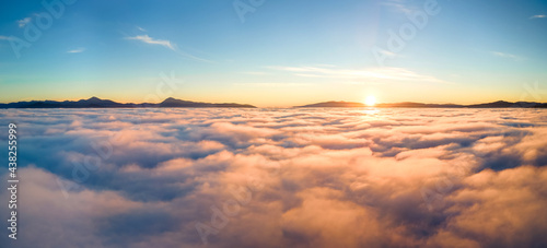 Aerial view of yellow sunset over white puffy clouds with distant mountains on horizon. © bilanol