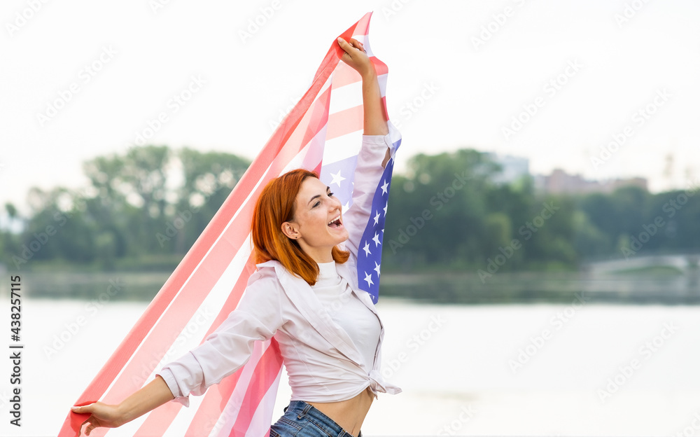 Portrait of happy smiling red haired girl with USA national flag in her hands. Positive young woman celebrating United States independence day.