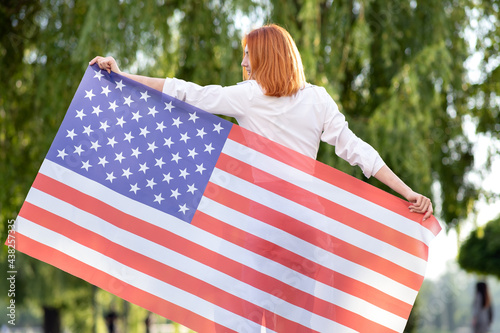 Rear view of happy young red haired woman posing with USA national flag standing outdoors in summer park. Positive girl celebrating United States independence day.