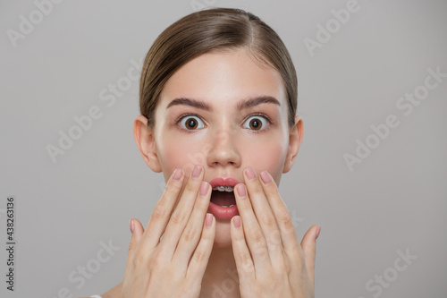 Beauty portrait. A beautiful girl with clean skin is surprised and covers her mouth with her hands. Gray background.