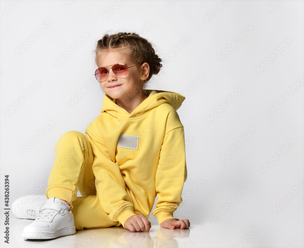Portrait of a cute stylish little girl sitting and looking at the camera on a white background. Child with a beautiful hairstyle is dressed in a yellow sports suit, sneakers and sunglasses. Portrait of a cute stylish little girl sitting and looking at the camera on a white background. Child with a beautiful hairstyle is dressed in a yellow sports suit, sneakers and sunglasses.