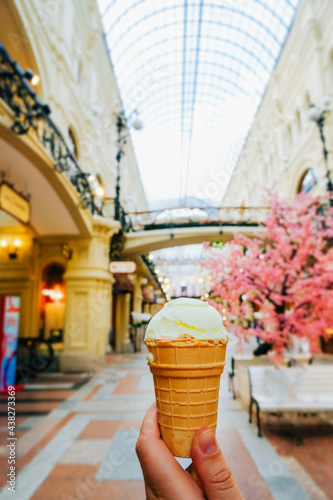Canvas Print hand holds creamy ice cream in waffle cup against the background of blurry shops and garlands in the mall