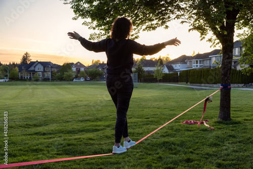 Adventurous White Caucasian Adult Woman walking on a Slackline between trees in a neighborhood park. Sunny Sunset. Surrey, Vancouver, British Columbia, Canada. photo