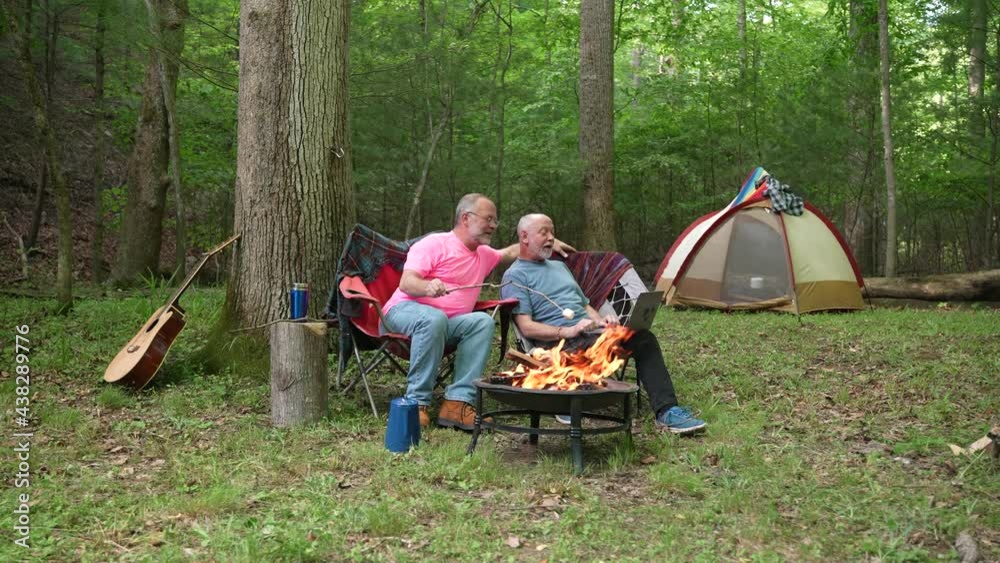 Two gay men with pride flag and tent in front of campfire talking to laptop on video chat. Two gay men with pride flag and tent in front of campfire talking to laptop on video chat.