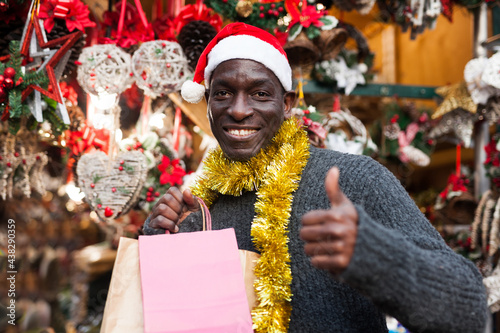 Portrait of smiling African American man in Santa hat with shopping bags on festive fair before Christmas.