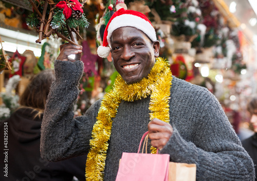 Smiling African American man in Santa hat selecting festive home decoration at outdoor Christmas fair..