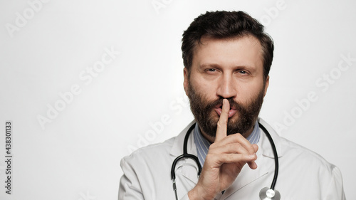Shh, doctor secret finger. Suspicious smiling man doctor on white background looking at camera and brings his index finger to his mouth lips and she say shhh