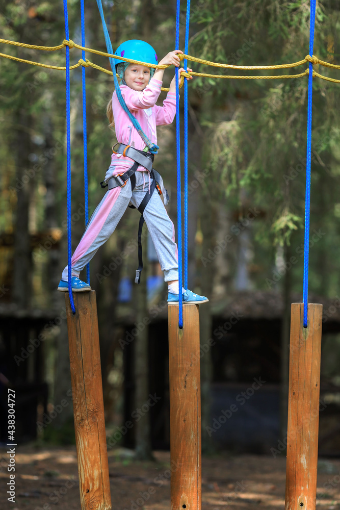 Fototapeta premium The child is engaged in sports.A girl overcomes sports obstacles in a rope park.A little girl in a protective helmet and tracksuit.Active recreation in the summer.Sports Rope Park for children