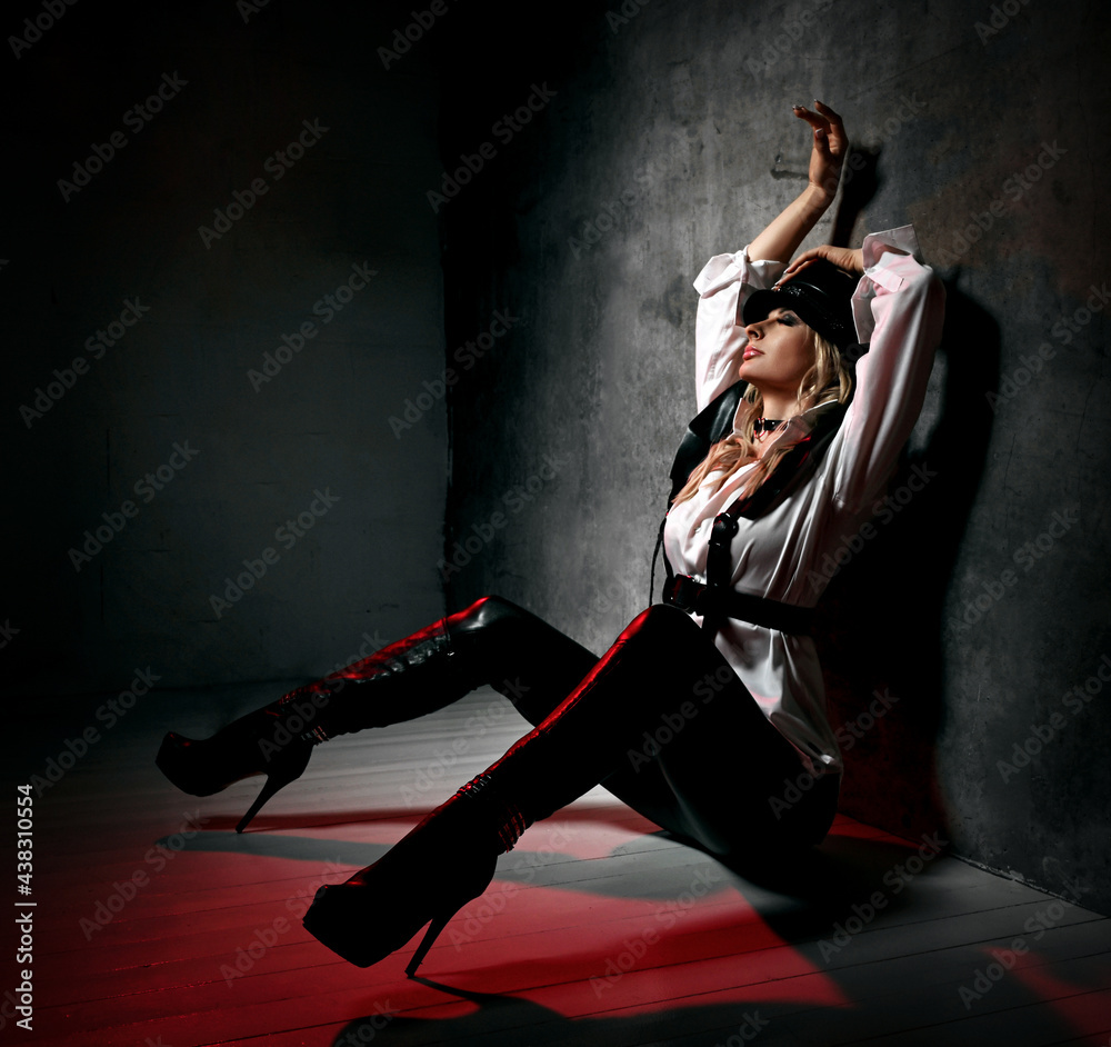 Sensual blonde woman in leather pants, white shirt and leather belts sits at concrete wall with her hands up, feeling pleasure and sexual arousal. Fashion, vogue, sexy stylish look for woman concept