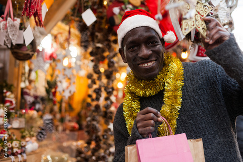 Cheerful African American in Santa hat looking for New Year decorations in traditional street market.