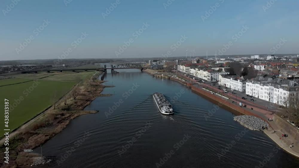 Following a large container cargo ship passing by embankment of historic medieval Hanseatic city of Zutphen, The Netherlands, besides river IJssel. Aerial urban transportation concept.
