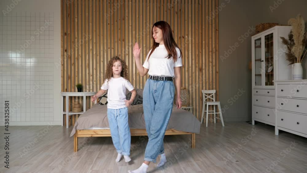 Joyful mother and little daughter in t-shirts and jeans dance and give high five with cheerful smiles, against large grey bed in bedroom at home Joyful mother and little daughter in t-shirts and jeans dance and give high five with cheerful smiles, against large grey bed in bedroom at home