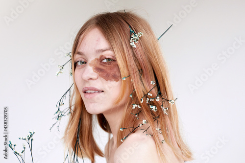 Positive young female model with brown spot on face skin and delicate flowers in hair smiling and looking at camera against white background photo