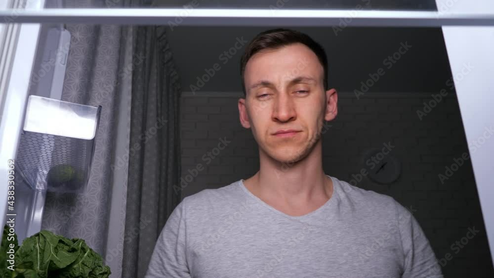Sleepy hungry man in grey t-shirt takes and bites delicious green apple and becomes happy in dark kitchen at night, close view from inside fridge Sleepy hungry man in grey t-shirt takes and bites delicious green apple and becomes happy in dark kitchen at night, close view from inside fridge