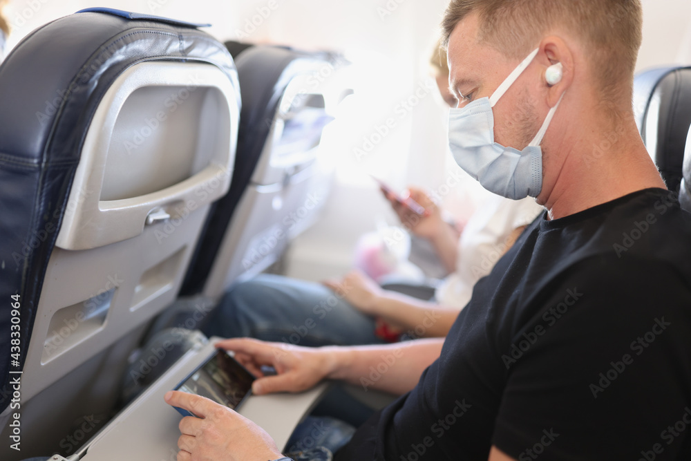 Man in medical protective mask sits on plane and watches video on his smartphone