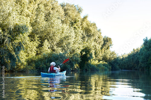 Woman row in blue kayak at wilderness areas of Danube river near green trees and thickets of wild grapes at spring