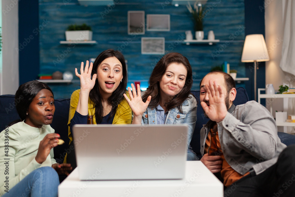 Obraz premium Happy multi-ethnic friends greeting her collegue during online videocall meeting using laptop webcam. Group of multiracial people spending time together on couch late at night during party