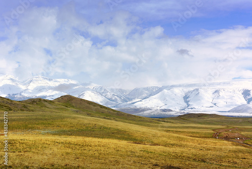 Kurai steppe in spring. Dry grass on the slopes, snow-capped peaks of the mountains of the Northern Chui range under a cloudy blue sky. Gorny Altai, Siberia, Russia
