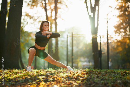 Fototapeta Naklejka Na Ścianę i Meble -  Attractive smiling woman with brown hair stretching legs during morning exercise at local park. Outdoor sport activity and regular training concept.